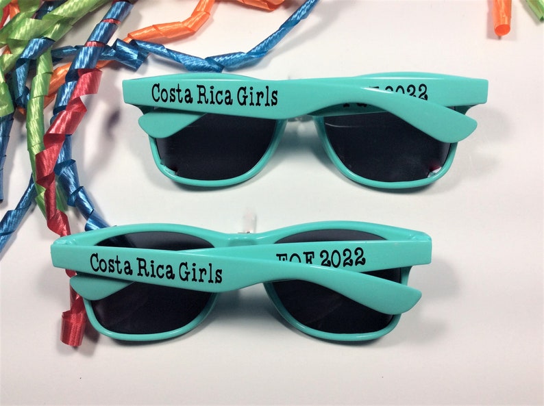 Op de afbeelding: Twee paar turquoise blauwe zonnebrillen met zwarte glazen. Het bovenste paar heeft de tekst "Costa Rica Girls" op het montuur. Het onderste paar heeft de tekst "TOF 2022" op het montuur.