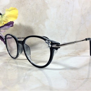 Puede incluir: Gafas con montura negra y lentes transparentes. Las monturas redondas est&aacute;n adornadas con peque&ntilde;os adornos brillantes cerca de las bisagras. Las patillas son plateadas y terminan en terminales negros. Fondo claro.