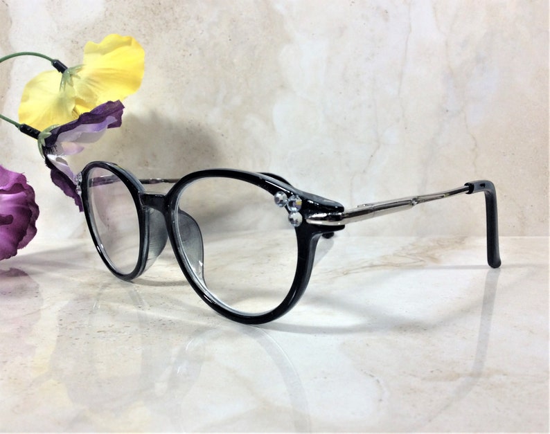 Puede incluir: Gafas con montura negra y lentes transparentes. Las monturas redondas est&aacute;n adornadas con peque&ntilde;os adornos brillantes cerca de las bisagras. Las patillas son plateadas y terminan en terminales negros. Fondo claro.