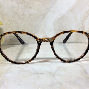 May include: Round eyeglasses with a tortoiseshell pattern frame. The frame is embellished with small, gold-colored rhinestones at the temples and bridge. The lenses are clear, and the arms are dark brown.