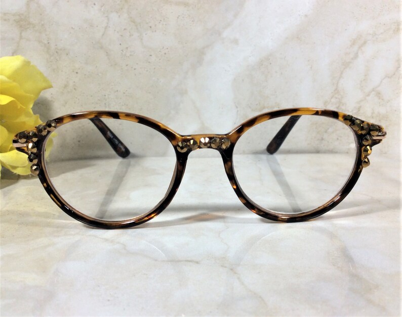May include: Round eyeglasses with a tortoiseshell pattern frame. The frame is embellished with small, gold-colored rhinestones at the temples and bridge. The lenses are clear, and the arms are dark brown.