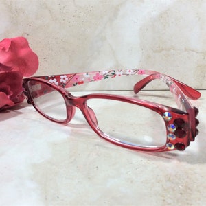 Puede incluir: Gafas con montura roja y lentes transparentes. Las monturas presentan un estampado floral en las varillas y pedrer&iacute;a decorativa en las esquinas. Una gran flor rosa en el fondo.