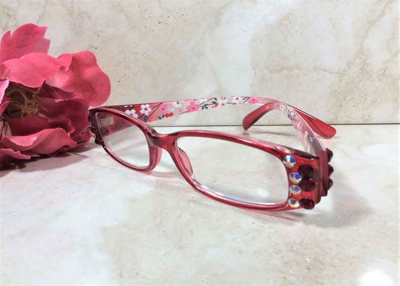 Puede incluir: Gafas con montura roja y lentes transparentes. Las monturas presentan un estampado floral en las varillas y pedrer&iacute;a decorativa en las esquinas. Una gran flor rosa en el fondo.