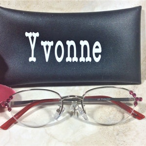 Pu&ograve; includere: Occhiali con montatura argentata, aste rosse e gemme rosa decorative. Gli occhiali sono davanti a un astuccio nero con il nome "Yvonne" a lettere bianche. Le lenti hanno protezione UV.