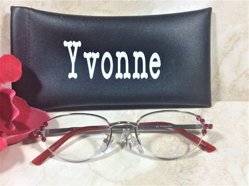 Pu&ograve; includere: Occhiali con montatura argentata, aste rosse e gemme rosa decorative. Gli occhiali sono davanti a un astuccio nero con il nome "Yvonne" a lettere bianche. Le lenti hanno protezione UV.