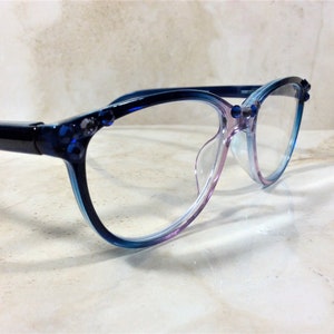 Puede incluir: Gafas con montura degradada azul y morada. Las lentes transparentes est&aacute;n engarzadas en una montura decorada con peque&ntilde;as joyas azules. Las gafas se exhiben sobre una superficie clara, con efecto m&aacute;rmol.