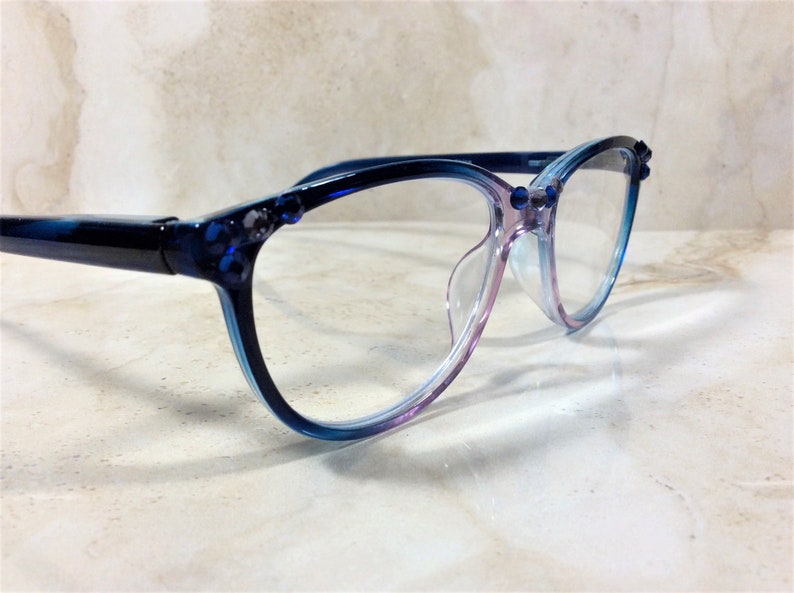 Puede incluir: Gafas con montura degradada azul y morada. Las lentes transparentes est&aacute;n engarzadas en una montura decorada con peque&ntilde;as joyas azules. Las gafas se exhiben sobre una superficie clara, con efecto m&aacute;rmol.