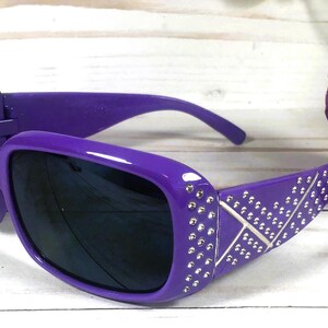 May include: Purple sunglasses with dark lenses. The frames are adorned with silver-colored rhinestones and a decorative X pattern. The sunglasses are set against a white background with a flower.