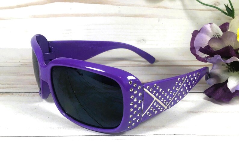 May include: Purple sunglasses with dark lenses. The frames are adorned with silver-colored rhinestones and a decorative X pattern. The sunglasses are set against a white background with a flower.