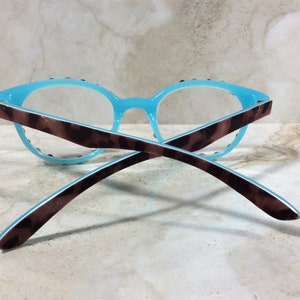 May include: A pair of eyeglasses with a light blue frame and clear lenses. The temples are brown with a tortoiseshell pattern. The glasses are resting on a light-colored surface.