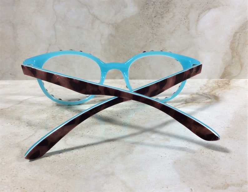 May include: A pair of eyeglasses with a light blue frame and clear lenses. The temples are brown with a tortoiseshell pattern. The glasses are resting on a light-colored surface.