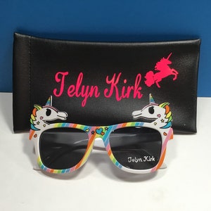 May include: Rainbow unicorn sunglasses with black lenses and a white frame. The sunglasses have a unicorn design on each side and the text "Jolyn Kirk" on the right lens. The sunglasses are in a black carrying case with the text "Jolyn Kirk" and a unicorn design.