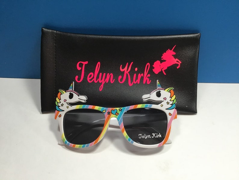May include: Rainbow unicorn sunglasses with black lenses and a white frame. The sunglasses have a unicorn design on each side and the text "Jolyn Kirk" on the right lens. The sunglasses are in a black carrying case with the text "Jolyn Kirk" and a unicorn design.