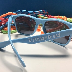 Peut inclure: Lunettes de soleil en plastique bleu clair avec des verres fonc&eacute;s. Les lunettes ont les mots "Birthday Squad" imprim&eacute;s sur le c&ocirc;t&eacute;.
