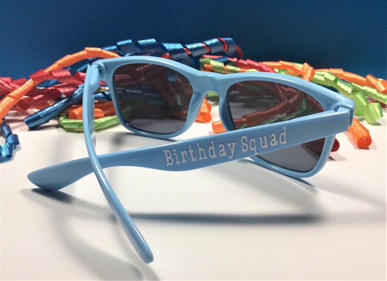 Peut inclure: Lunettes de soleil en plastique bleu clair avec des verres fonc&eacute;s. Les lunettes ont les mots "Birthday Squad" imprim&eacute;s sur le c&ocirc;t&eacute;.