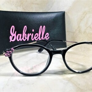 May include: Black-framed eyeglasses with clear lenses and pink jewel accents on the corners. The glasses are paired with a black case with the name "Gabrielle" in pink script. The glasses have a modern, stylish design.