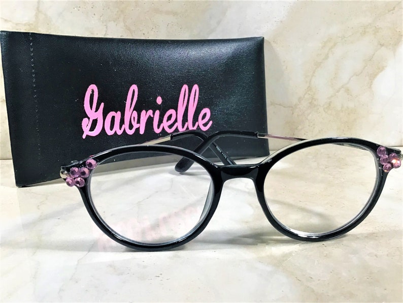 May include: Black-framed eyeglasses with clear lenses and pink jewel accents on the corners. The glasses are paired with a black case with the name "Gabrielle" in pink script. The glasses have a modern, stylish design.