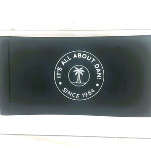 May include: Black zippered pouch with a white circle logo that says "It's All About Dan! Since 1964" and a palm tree inside the circle.