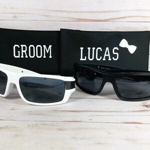 May include: Two pairs of sunglasses, one white and one black, rest on a light wood surface. The white sunglasses have a thick frame. Behind them are black cases with the words "GROOM" and "LUCAS" printed in white.