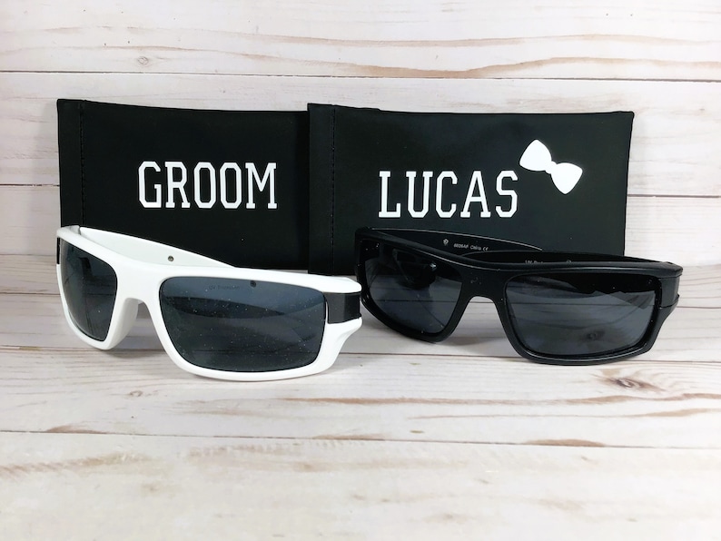 May include: Two pairs of sunglasses, one white and one black, rest on a light wood surface. The white sunglasses have a thick frame. Behind them are black cases with the words "GROOM" and "LUCAS" printed in white.