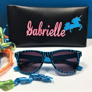 May include: A black zippered case with the name "Gabrielle" printed in pink letters and a blue unicorn silhouette. The case is open and a pair of blue and black zebra-striped sunglasses are sitting on a white surface.