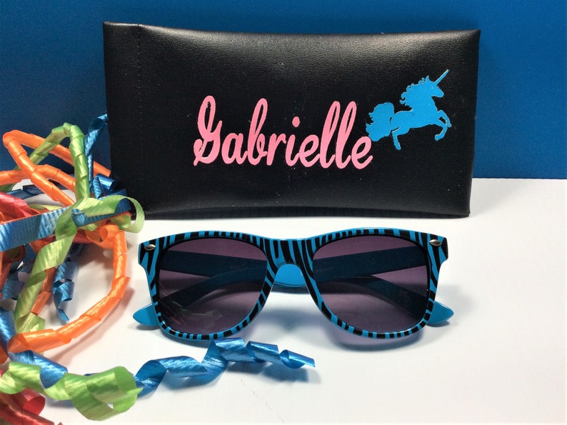 May include: A black zippered case with the name "Gabrielle" printed in pink letters and a blue unicorn silhouette. The case is open and a pair of blue and black zebra-striped sunglasses are sitting on a white surface.