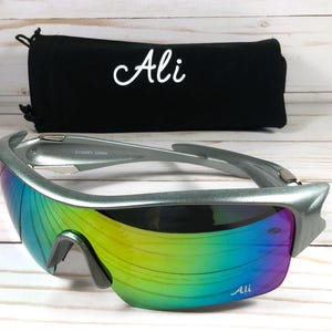 May include: Silver framed sunglasses with a rainbow iridescent lens. The sunglasses have the word "Ali" printed on the frame and the lens. The sunglasses are sitting on a wooden surface.
