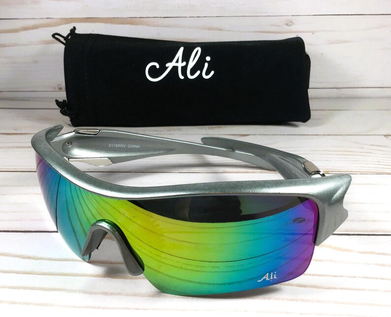 May include: Silver framed sunglasses with a rainbow iridescent lens. The sunglasses have the word "Ali" printed on the frame and the lens. The sunglasses are sitting on a wooden surface.