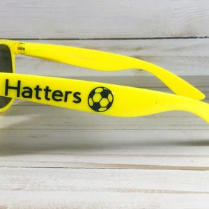 Puede incluir: Gafas de sol de color amarillo brillante con lentes oscuras. La patilla lateral muestra el texto negro "Hatters" seguido de un gr&aacute;fico de un bal&oacute;n de f&uacute;tbol en blanco y negro. La montura est&aacute; fabricada en pl&aacute;stico amarillo s&oacute;lido con acabado liso.