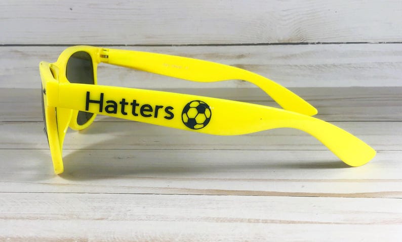 Puede incluir: Gafas de sol de color amarillo brillante con lentes oscuras. La patilla lateral muestra el texto negro "Hatters" seguido de un gr&aacute;fico de un bal&oacute;n de f&uacute;tbol en blanco y negro. La montura est&aacute; fabricada en pl&aacute;stico amarillo s&oacute;lido con acabado liso.