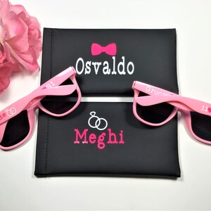 May include: Two pairs of pink sunglasses with black lenses. The sunglasses are personalized with the words "Bride" and "Groom" and the names "Meghi" and "Osvaldo".
