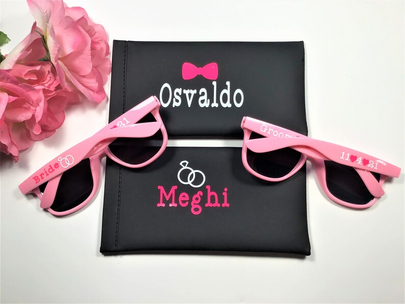 May include: Two pairs of pink sunglasses with black lenses. The sunglasses are personalized with the words "Bride" and "Groom" and the names "Meghi" and "Osvaldo".