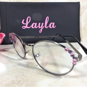 Puede incluir: Gafas de forma ovalada con montura de metal oscuro y lentes transparentes. La montura est&aacute; adornada con peque&ntilde;as piedras preciosas moradas. Una funda negra con el nombre "Layla" en escritura rosa est&aacute; detr&aacute;s de las gafas. Una flor rosa est&aacute; en el fondo.