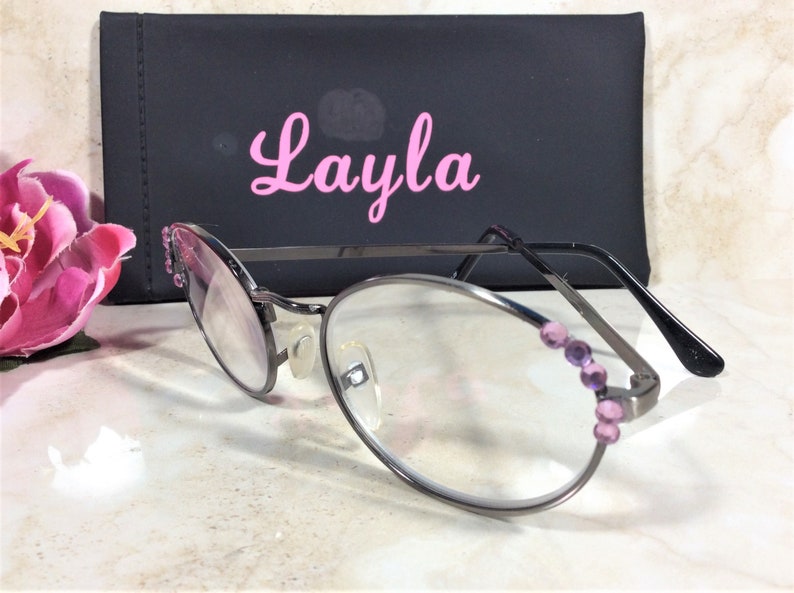 Puede incluir: Gafas de forma ovalada con montura de metal oscuro y lentes transparentes. La montura est&aacute; adornada con peque&ntilde;as piedras preciosas moradas. Una funda negra con el nombre "Layla" en escritura rosa est&aacute; detr&aacute;s de las gafas. Una flor rosa est&aacute; en el fondo.