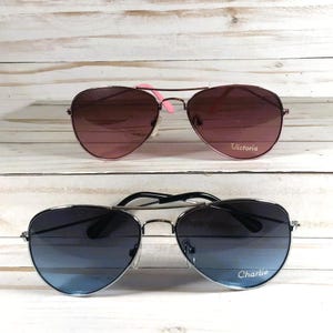 May include: Two pairs of aviator sunglasses. The top pair features pink-tinted lenses and pink temple tips with the name Victoria printed on the right lens. The bottom pair has blue-tinted lenses and black temple tips with the name Charlie printed.
