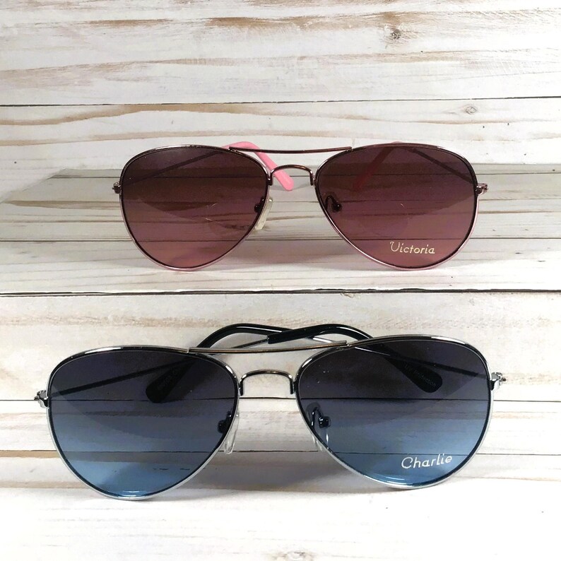 May include: Two pairs of aviator sunglasses. The top pair features pink-tinted lenses and pink temple tips with the name Victoria printed on the right lens. The bottom pair has blue-tinted lenses and black temple tips with the name Charlie printed.