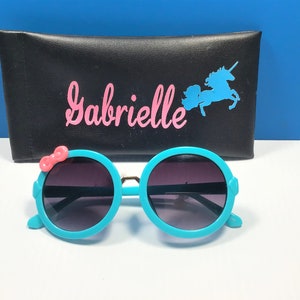 May include: Teal round sunglasses with a pink bow on the side. The sunglasses are in front of a black case with the name "Gabrielle" and a unicorn silhouette.