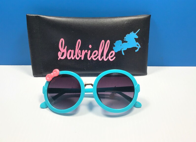 May include: Teal round sunglasses with a pink bow on the side. The sunglasses are in front of a black case with the name "Gabrielle" and a unicorn silhouette.