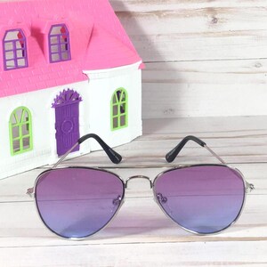 Peut inclure: Lunettes de soleil aviateur argent&eacute;es avec des verres d&eacute;grad&eacute;s violets. Les lunettes de soleil sont sur une surface en bois blanc.