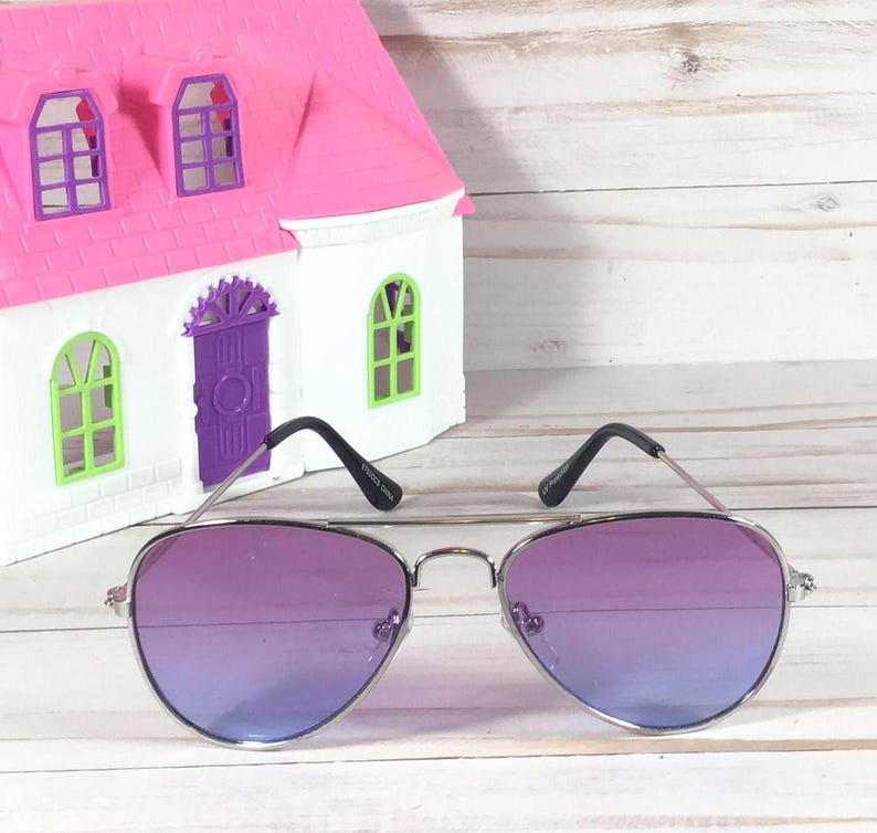 Peut inclure: Lunettes de soleil aviateur argent&eacute;es avec des verres d&eacute;grad&eacute;s violets. Les lunettes de soleil sont sur une surface en bois blanc.