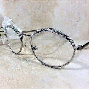 May include: Silver-framed eyeglasses with clear lenses. The top rims are embellished with a row of sparkling, clear rhinestones. The temples are black, and the glasses have nose pads. A sprig of white flowers is in the background.