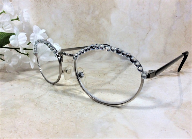May include: Silver-framed eyeglasses with clear lenses. The top rims are embellished with a row of sparkling, clear rhinestones. The temples are black, and the glasses have nose pads. A sprig of white flowers is in the background.
