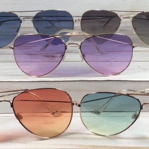 May include: Six pairs of aviator sunglasses with gradient lenses in various colours, including blue, purple, orange, and green. The sunglasses have gold-coloured frames and are displayed on a white wooden surface.