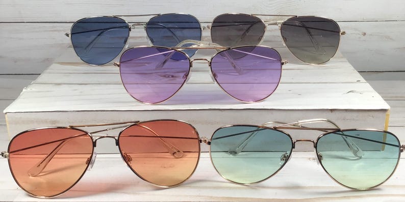 May include: Six pairs of aviator sunglasses with gradient lenses in various colours, including blue, purple, orange, and green. The sunglasses have gold-coloured frames and are displayed on a white wooden surface.