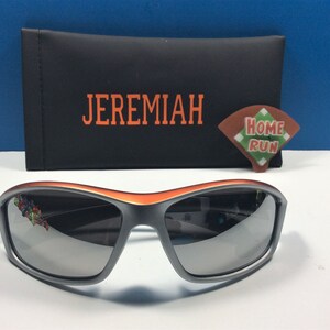 Puede incluir: Gafas de sol grises con un borde naranja y lentes espejados. Las gafas de sol est&aacute;n en una caja negra con la palabra "JEREMIAH" impresa en naranja. Una peque&ntilde;a pegatina naranja y verde "HOME RUN" est&aacute; en la caja.