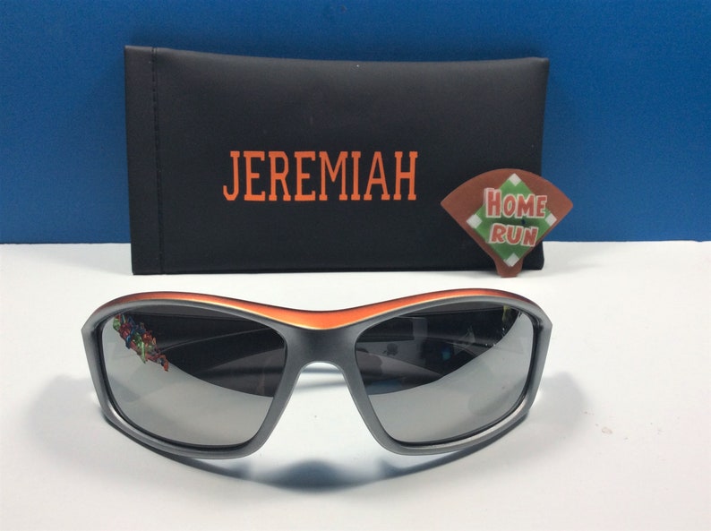 Puede incluir: Gafas de sol grises con un borde naranja y lentes espejados. Las gafas de sol est&aacute;n en una caja negra con la palabra "JEREMIAH" impresa en naranja. Una peque&ntilde;a pegatina naranja y verde "HOME RUN" est&aacute; en la caja.