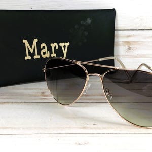 May include: Gold-framed aviator sunglasses with gradient lenses sit on a light-coloured wooden surface. A black case with the name "Mary" in gold lettering is behind the sunglasses. The sunglasses have a classic teardrop shape.