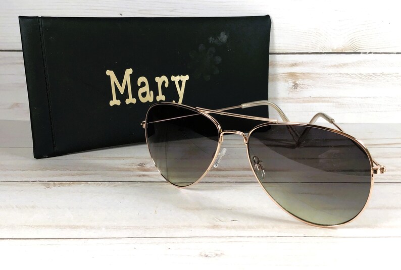 May include: Gold-framed aviator sunglasses with gradient lenses sit on a light-coloured wooden surface. A black case with the name "Mary" in gold lettering is behind the sunglasses. The sunglasses have a classic teardrop shape.
