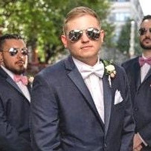 May include: Four men wearing navy blue suits with white shirts and pink bow ties. The men are wearing sunglasses and are standing in a row. The man in the center is wearing a white bow tie.