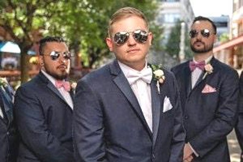 May include: Four men wearing navy blue suits with white shirts and pink bow ties. The men are wearing sunglasses and are standing in a row. The man in the center is wearing a white bow tie.